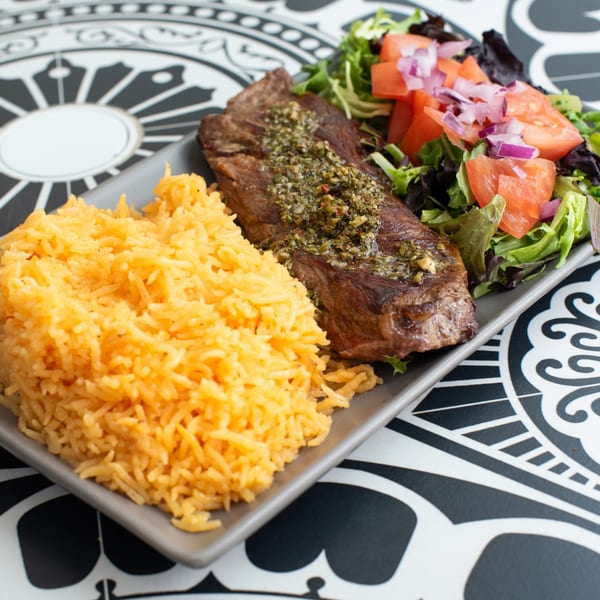 Grilled steak with chimichurri, yellow rice, and a side salad with tomatoes and red onions.
