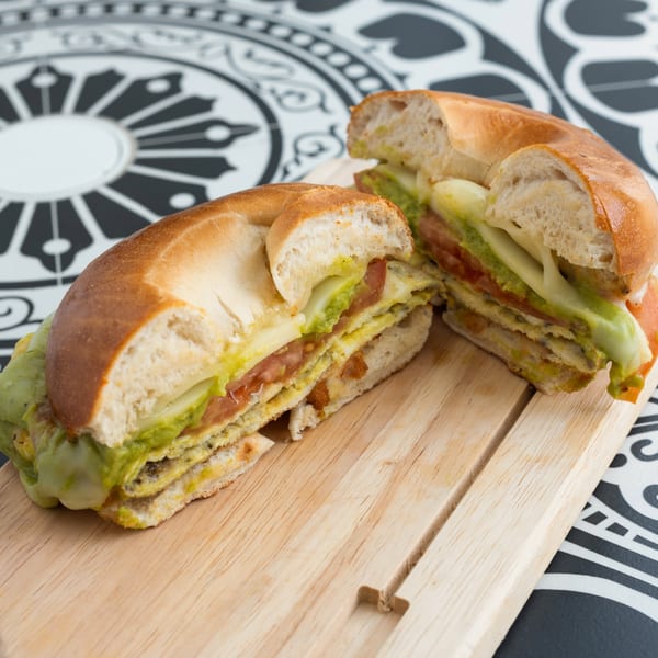 A sliced bagel sandwich with avocado, cheese, and tomato on a wooden board.