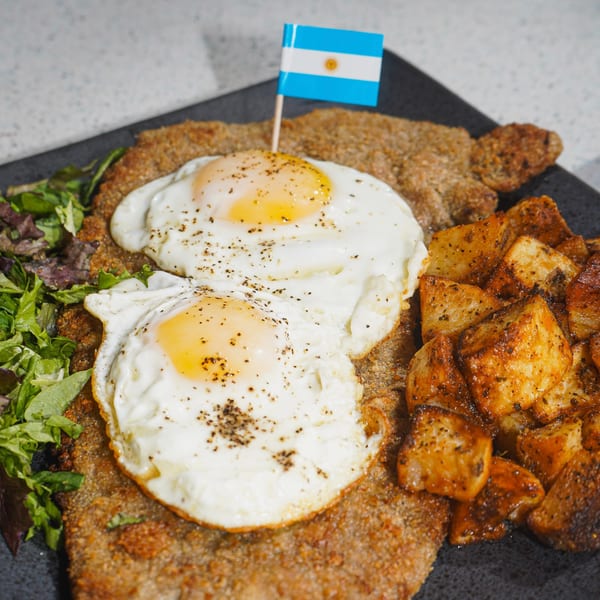 Milanesa a caballo with two fried eggs, roasted potatoes, and a side salad, garnished with an Argentinian flag.