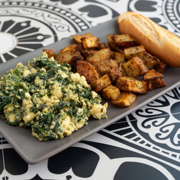 Creamy green spinach and cheese scramble with roasted potatoes and a baguette on a gray plate.